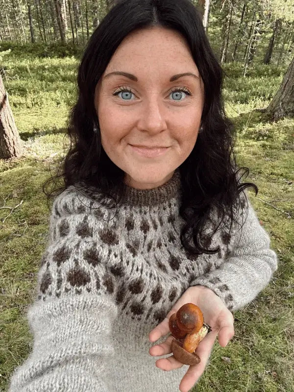 A woman with long dark hair and blue eyes wears the Sisu Knitwear Terhoneule neuleohje Suomi PDF sweater, featuring a patterned pyöreä kaarroke, as she smiles in a forest holding a brown mushroom.