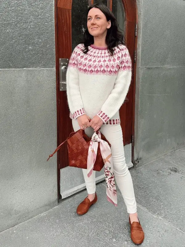 A woman stands in front of a metal door wearing white pants, brown loafers, holding a brown woven bag with a pink scarf, and the Sisu Knitwear Vadelmaneule neuleohje Suomeksi PDF sweater in white and pink pattern.