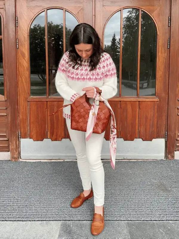 A woman wearing the Sisu Knitwear Vadelmaneule neuleohje Suomeksi PDF white and pink patterned sweater, white pants, and brown shoes stands on a mat by wooden doors, looking down at her brown woven handbag with a pink scarf tied to it.