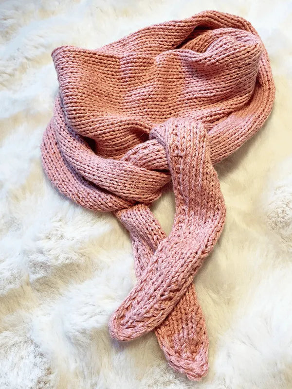 A cozy, pink knitted scarf made from Knitting for Olive yarn is loosely coiled on a fluffy white surface. The design is created using the Somany huivi ohje PDF pattern, highlighting its soft and inviting texture.
