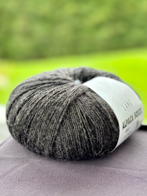 A close-up of Lang Alpaca Soxx 4ply dark gray yarn with a white label, perfect for knitting with the Mittums lapasten ohje (PDF). The ball rests on fabric, set against a softly blurred green background for a tranquil feel.