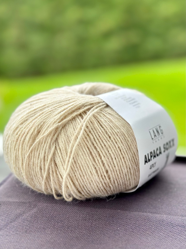 A ball of Lang Alpaca Soxx 4ply in light beige with a white label and the Mittums lapasten ohje (PDF) rests on a gray surface, set against a blurred green outdoor background.