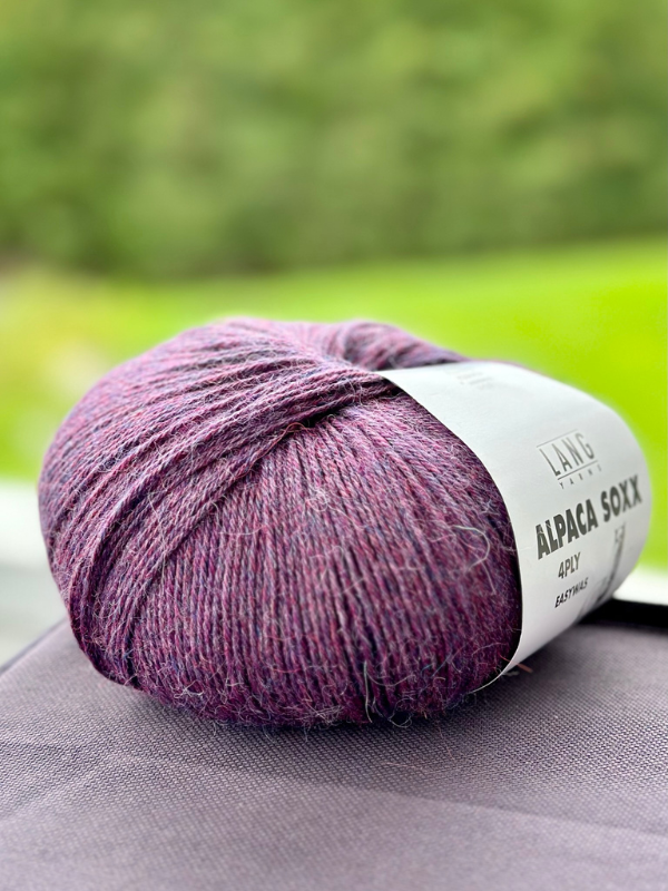 A ball of Lang Alpaca Soxx 4ply yarn and the Mittums lapasten ohje (PDF) pattern, ideal for tumput, sits on a surface against a blurred green background.