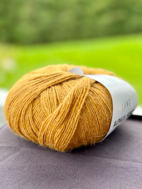 A close-up of Lang Alpaca Soxx 4ply yarn in mustard yellow with a white label rests on dark fabric, evoking outdoor inspiration—perfect for creating the Mittums lapasten ohje (PDF) pattern.