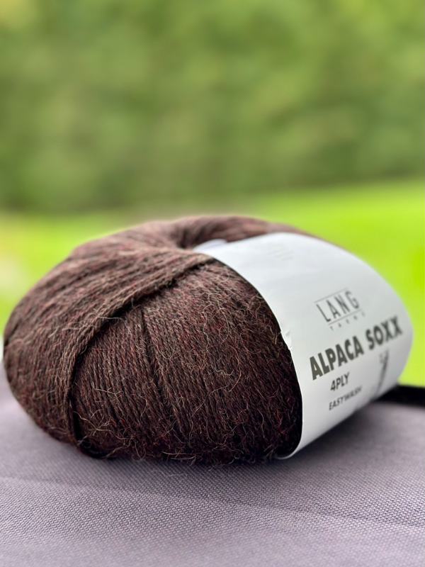 A ball of dark brown Lang Alpaca Soxx 4ply sits on a table with a blurred green outdoor background, featured in the Mittums lapasten ohje (PDF) + Lang Alpaca Soxx 4ply set—perfect for your next knitting project.