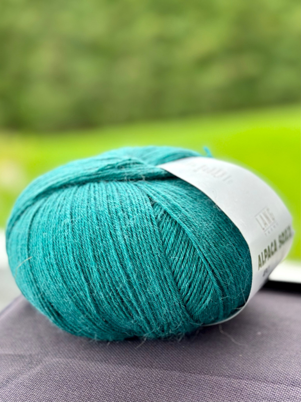 A close-up of a teal Lang Alpaca Soxx 4ply yarn ball with a white label on a dark surface, set against a blurred green outdoor background—ideal for Mittums lapasten ohje (PDF) mitten projects.