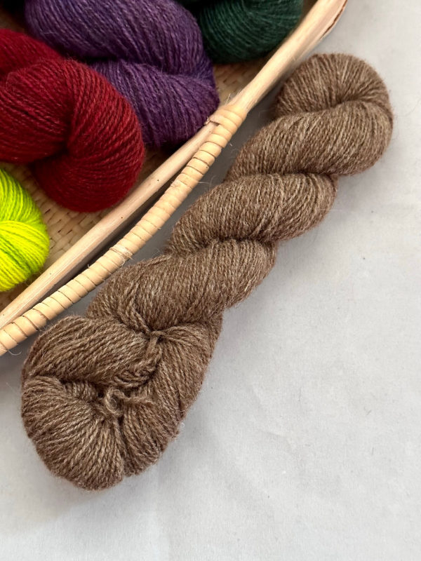 A skein of brown WoolWomen Huiku yarn rests on a light surface beside a basket filled with red, purple, green, and yellow skeins.