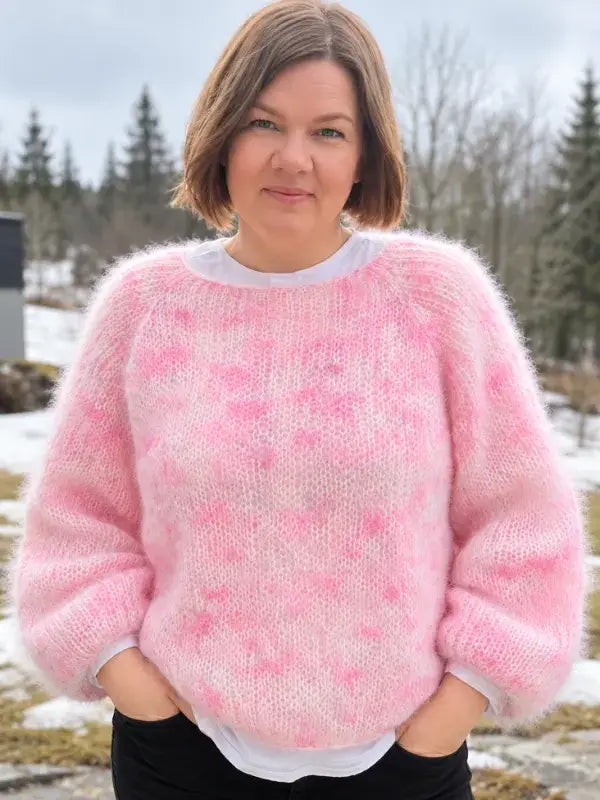 A woman with short brown hair stands outdoors, wearing a fluffy pink sweater made using the Hattara neuleohje pattern over a white shirt. Snow and trees are visible in the background. She has her hands in her pockets and is looking at the camera.