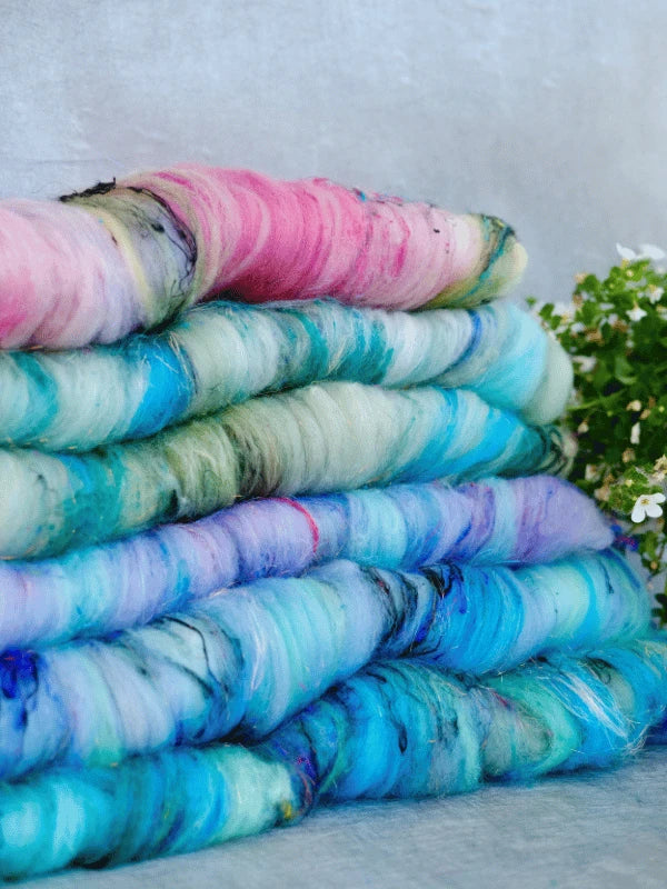 Four stacked WoolWomen Art Batt -Cormo batts of käsinvärjätty villa in pastel pink, blue, and green rest on a gray surface, with a small green plant with white flowers in the background. Great for kehräyskuitu projects.