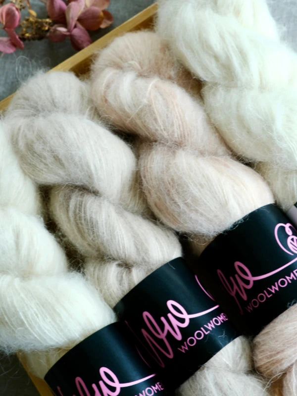 Four skeins of soft, fluffy WoolWomen Alpaca Glimmer yarn in off-white and pale peach hues lie side by side, each elegantly labeled. Spun from luxurious baby alpaca and meticulously hand-dyed as käsinvärjätty alpakkalanka, these skeins exude warmth and sophistication. Delicate pink flowers can be seen partially in the background.
