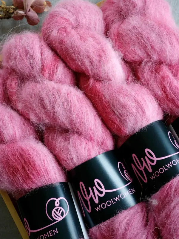Three skeins of fluffy pink WoolWomen Alpaca Glimmer yarn, each with a black label displaying the Wool Women name and a stylized logo, lying closely together on a flat surface.