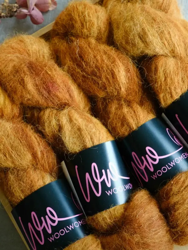 Four skeins of golden brown WoolWomen Alpaca Glimmer yarn are wrapped with black labels that feature the brand name in pink and white lettering. A dried flower is partially visible in the upper left corner.