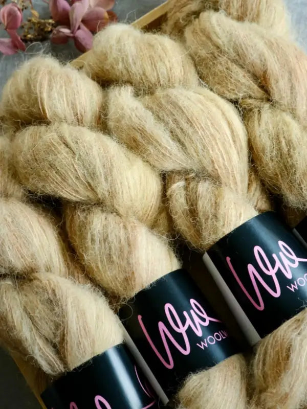 Three skeins of light tan, fluffy WoolWomen Alpaca Glimmer yarn with black labels featuring pink writing. There are also some pink flowers in the background.