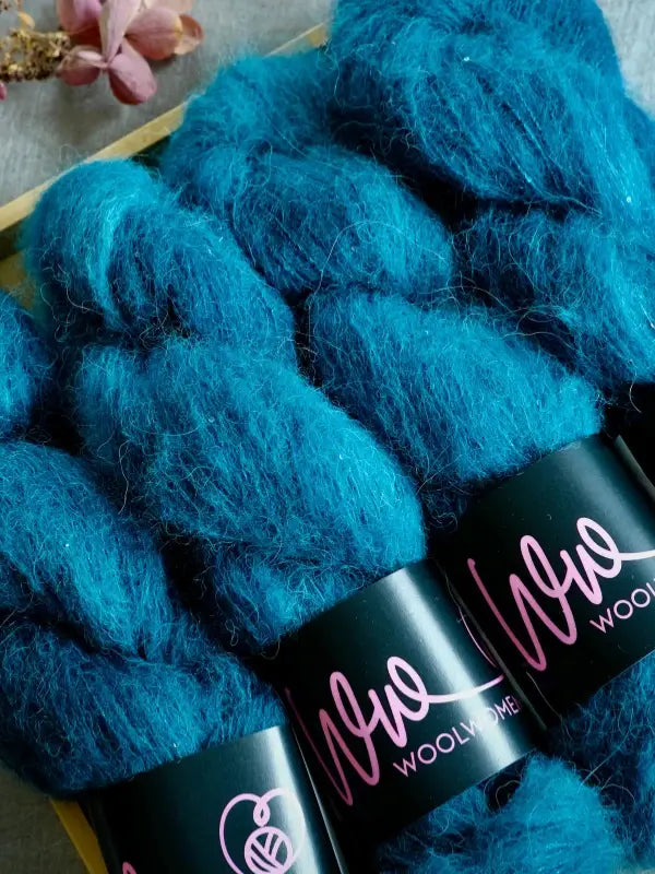 Three bundles of fluffy, teal blue WoolWomen Alpaca Glimmer yarn with black labels reading “Wool” lie side by side on a wooden surface, with a pink flower slightly visible in the upper left corner.
