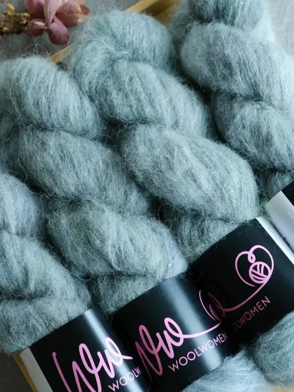 Skeins of soft, pale blue-green yarn labeled WoolWomen Alpaca Glimmer—crafted from luxurious baby alpaca and käsinvärjätty alpakkalanka—are arranged closely together on a light wooden surface, with a small bundle of dried flowers in the corner.