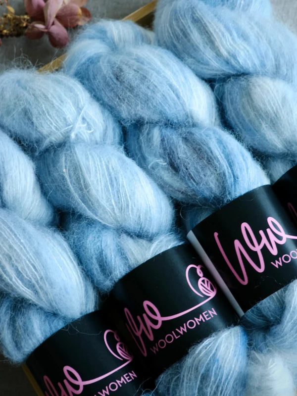 Three skeins of WoolWomen Alpaca Glimmer, a soft, light blue baby alpaca yarn with a fluffy texture. Each skein is wrapped in a black label featuring “Wool Women” in pink script. A dried flower rests in the corner.