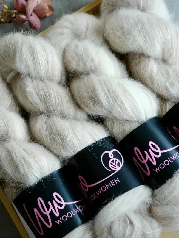 Three skeins of soft, cream-colored WoolWomen Alpaca Glimmer yarn with a pink logo are arranged closely together. Dried pink flowers can be seen in the upper left corner of the image.