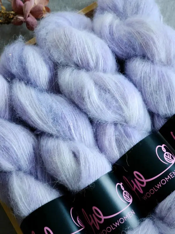 Three skeins of soft, fluffy lavender-colored WoolWomen Alpaca Glimmer yarn are arranged side by side, each wrapped with a black label featuring pink writing. Dried flowers are placed in the background.
