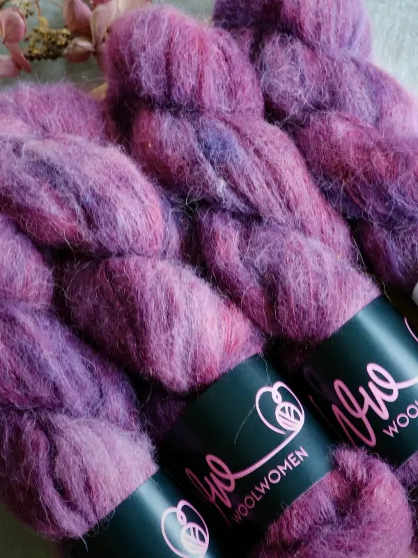 Three skeins of fluffy, purple and pink variegated WoolWomen Alpaca Glimmer yarn with sleek black labels featuring “WoolWomen” in bright pink lettering. This luxurious käsinvärjätty (hand-dyed) alpakkalanka showcases stunning color blends, while a softly blurred pink flower appears in the background.