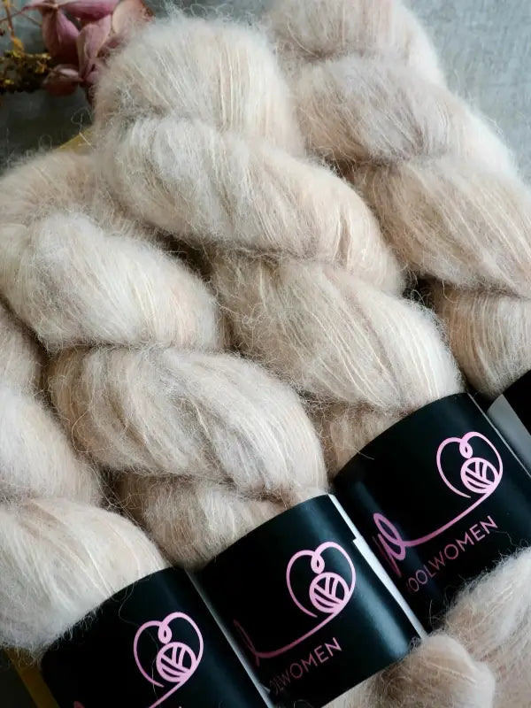Four skeins of soft, fluffy, pale beige WoolWomen Alpaca Glimmer yarn are bundled together, each with a black label featuring a pink yarn ball logo and the word WOOLWOMEN. A dried flower is partially visible in the background.