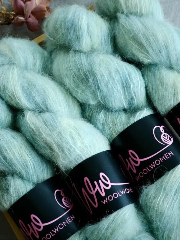 Three skeins of soft, light teal WoolWomen Alpaca Glimmer yarn with black and pink wrappers are arranged together on a surface, with a dried flower in the background.