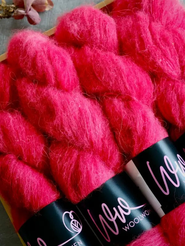 Three skeins of bright pink WoolWomen Alpaca Glimmer are arranged side by side in a box, each with a black label that reads “Wool” in cursive pink lettering. A pink flower is visible in the upper left corner.