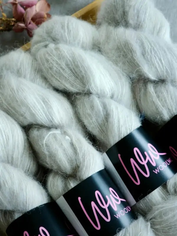 Three skeins of WoolWomen Alpaca Glimmer, a soft, light gray käsinvärjätty alpakkalanka, each wrapped with a black label featuring ‘Woo’ in pink text, arranged side by side on a wooden surface with dried pink flowers in the background.
