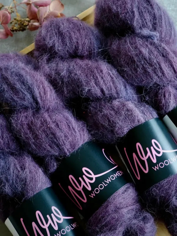 Four skeins of WoolWomen Alpaca Glimmer—luxuriously fluffy purple yarn made from käsinvärjätty alpakkalanka—are arranged on a surface, with dried flowers visible in the background.