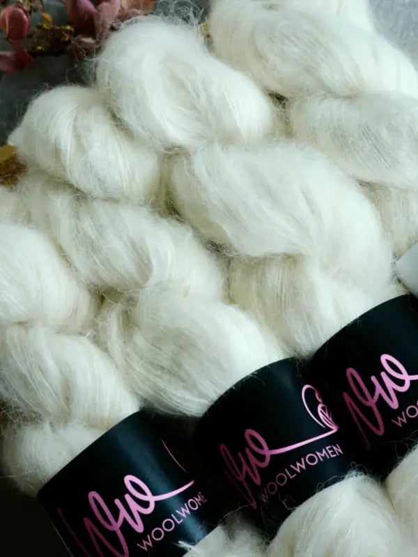 Three skeins of soft, white WoolWomen Alpaca Glimmer are braided and bundled together, each labeled with a black band featuring pink cursive text that reads Wool Women. This käsinvärjätty alpakkalanka offers a luxurious feel and beautiful finish, perfect for your next knitting project.