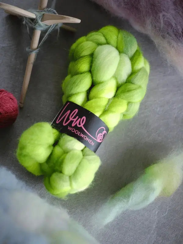 Two skeins of käsinvärjätty WoolWomen kehruukuitu Corriedale in bright green rest on a gray surface, with a white drop spindle and tuft of wool nearby—ideal for kehräämiseen.