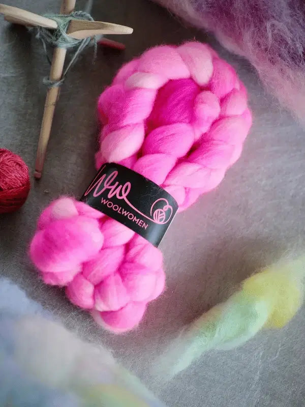 Two bundles of WoolWomen kehruukuitu Corriedale käsinvärjätty, thick braided pink wool with black WoolWomen labels, rest on a gray surface surrounded by other wool fibers and a white wooden star.