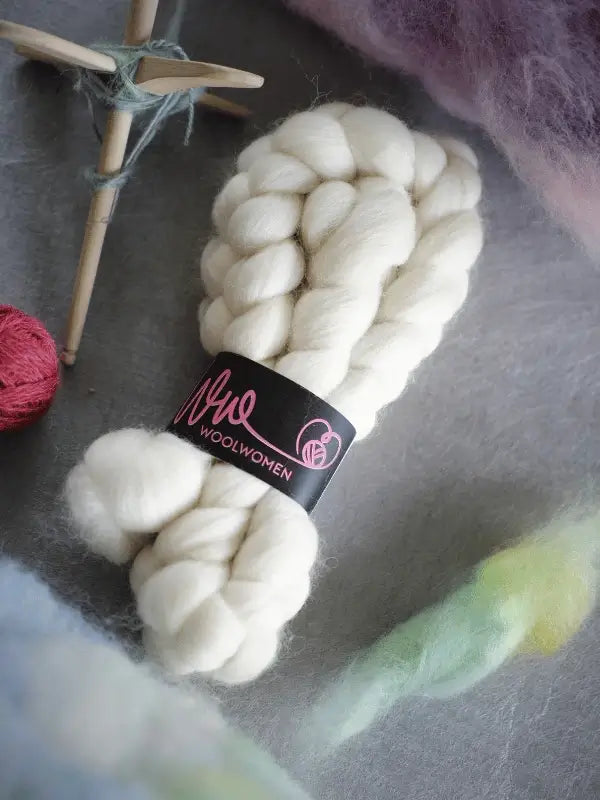 Two braids of WoolWomen kehruukuitu Corriedale Natural are on a gray surface, surrounded by green spinning fiber tufts and a wooden drop spindle with partially spun yarn.