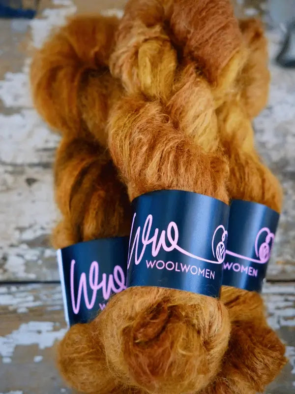 Three skeins of WoolWomen Fluff Mohair yarn in golden brown, each with a black label and pink logo, are displayed on a rustic, weathered wooden surface.