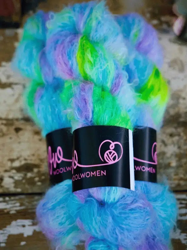 Two skeins of WoolWomen Fluff Mohair in blue, green, and purple hues with pink WOOLWOMEN labels, displayed on a rustic wooden surface.