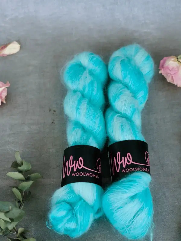Two skeins of bright turquoise WoolWomen Silk Mohair with black labels lie side by side on a gray surface, surrounded by small pink flowers and green foliage.
