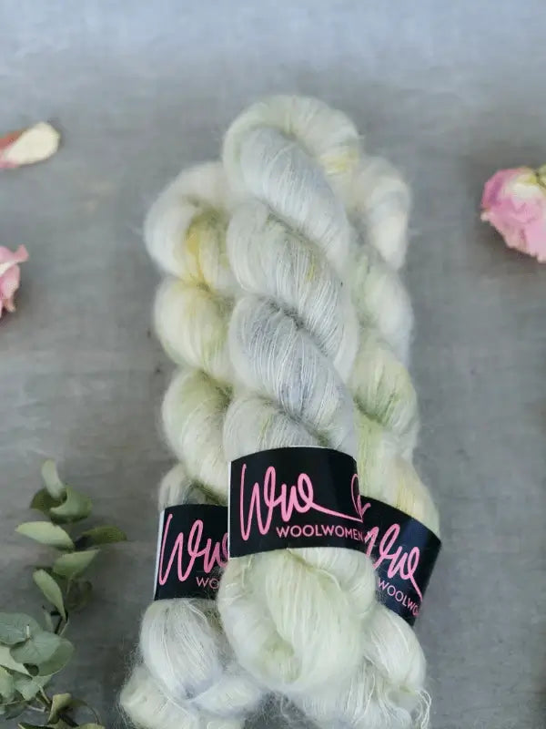 Two skeins of soft, pale green and white WoolWomen Silk Mohair yarn with labels rest on a gray surface, surrounded by dried flowers and greenery.
