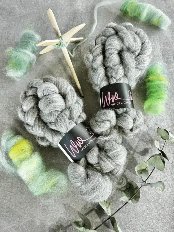 Two bundles of WoolWomen Kehruuvilla BFL Natural are displayed with green and yellow spinning fiber, a wooden drop spindle, and a dried branch on a gray surface.
