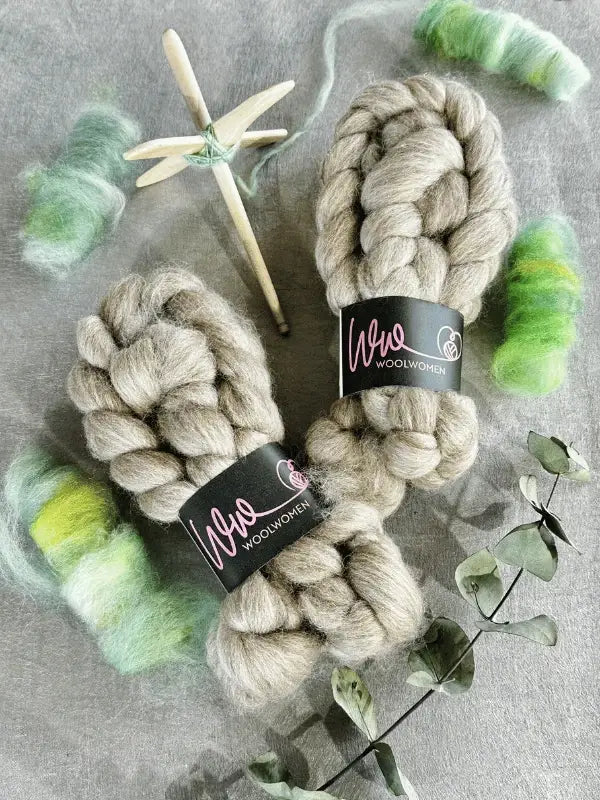 Two braids of gray WoolWomen Kehruuvilla BFL Natural wool, with green and yellow Bluefaced Leicester fiber, a wooden drop spindle wound with yarn, and eucalyptus leaves on a light surface.