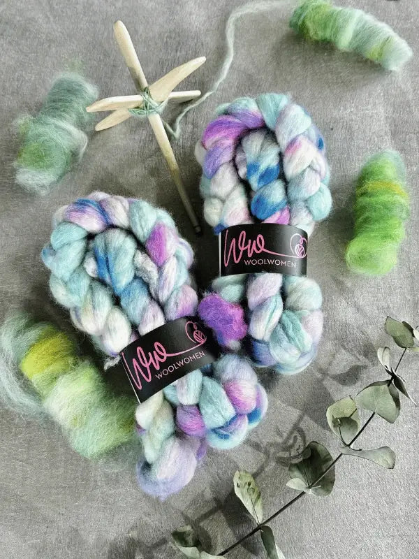 Two skeins of chunky, multicolored WoolWomen Kehruuvilla BFL Käsinvärjätyt rest on a gray surface, surrounded by hand-dyed spinning fiber in green and blue shades, a wooden spindle, and eucalyptus branches.