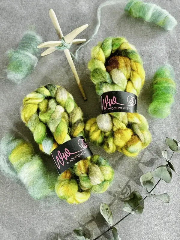 Two skeins of green, yellow, and brown WoolWomen Kehruuvilla BFL Käsinvärjätyt rest on a gray surface, surrounded by rolled wool, a spindle, and dried eucalyptus leaves.