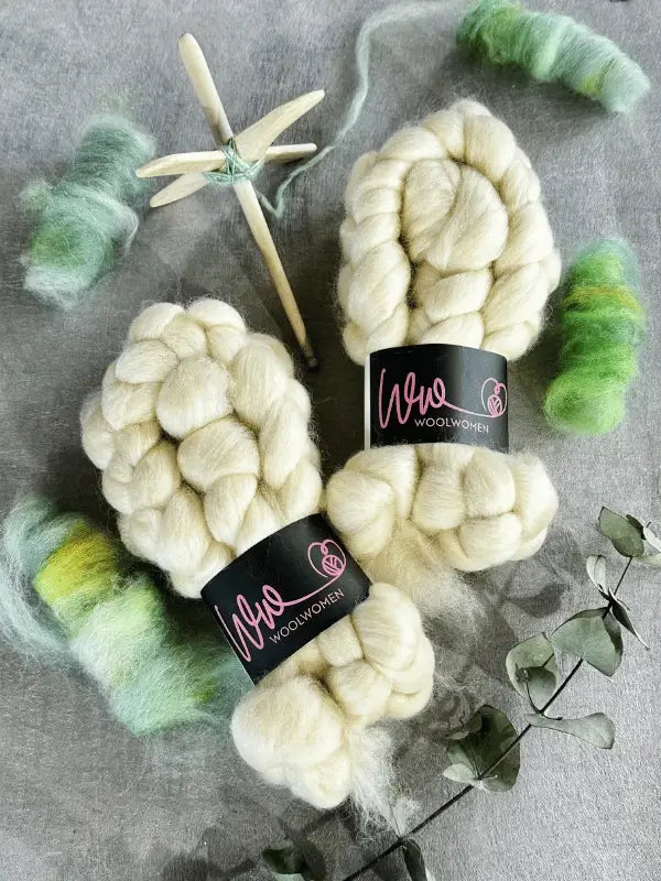 Two braids of WoolWomen Kehruuvilla BFL Natural, a wooden drop spindle, and green-yellow batts are arranged on a gray surface with eucalyptus leaves nearby.
