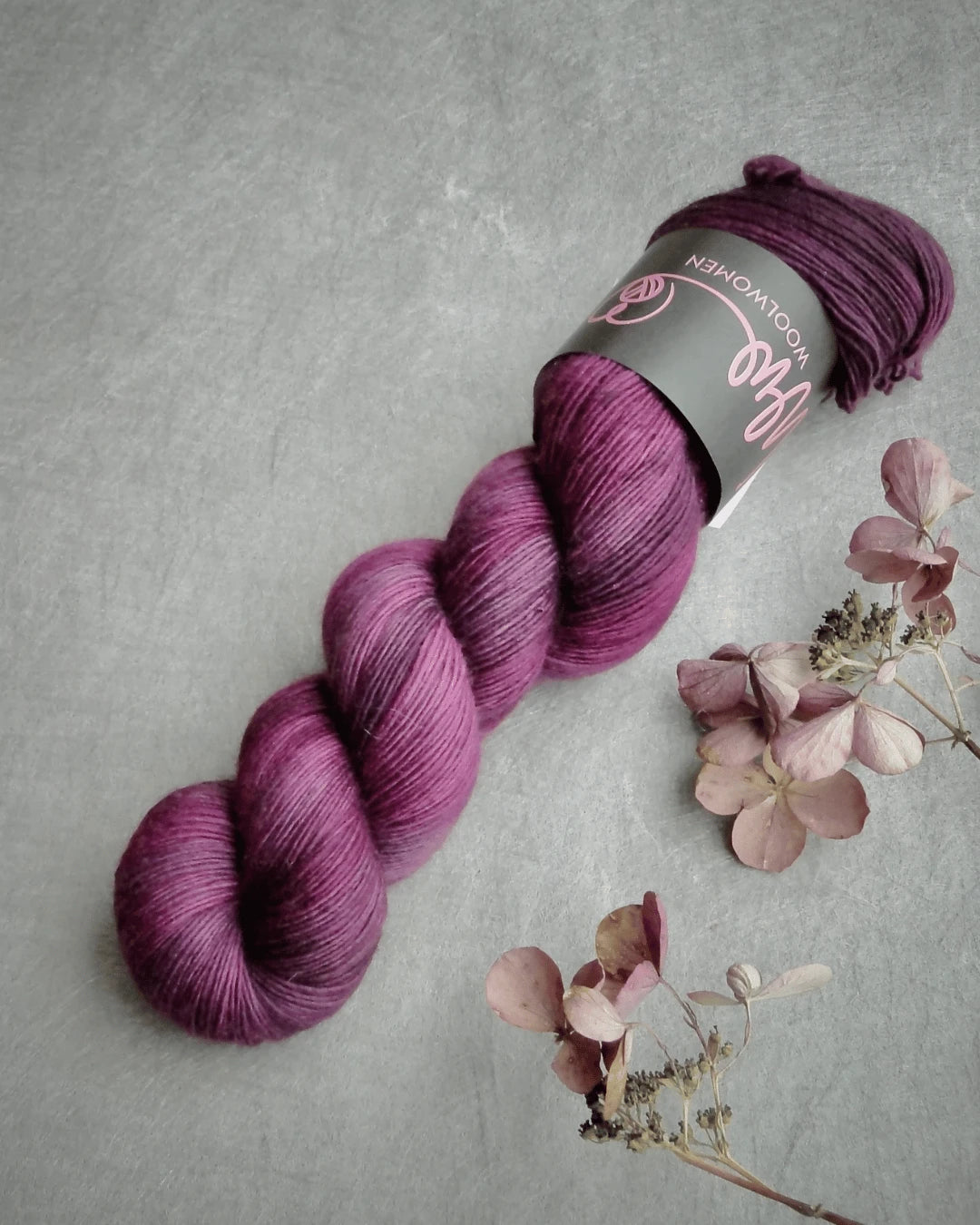 A skein of WoolWomen Merino Single käsinvärjätty merinovilla purple yarn with a black label rests on a gray surface next to dried hydrangea flowers.