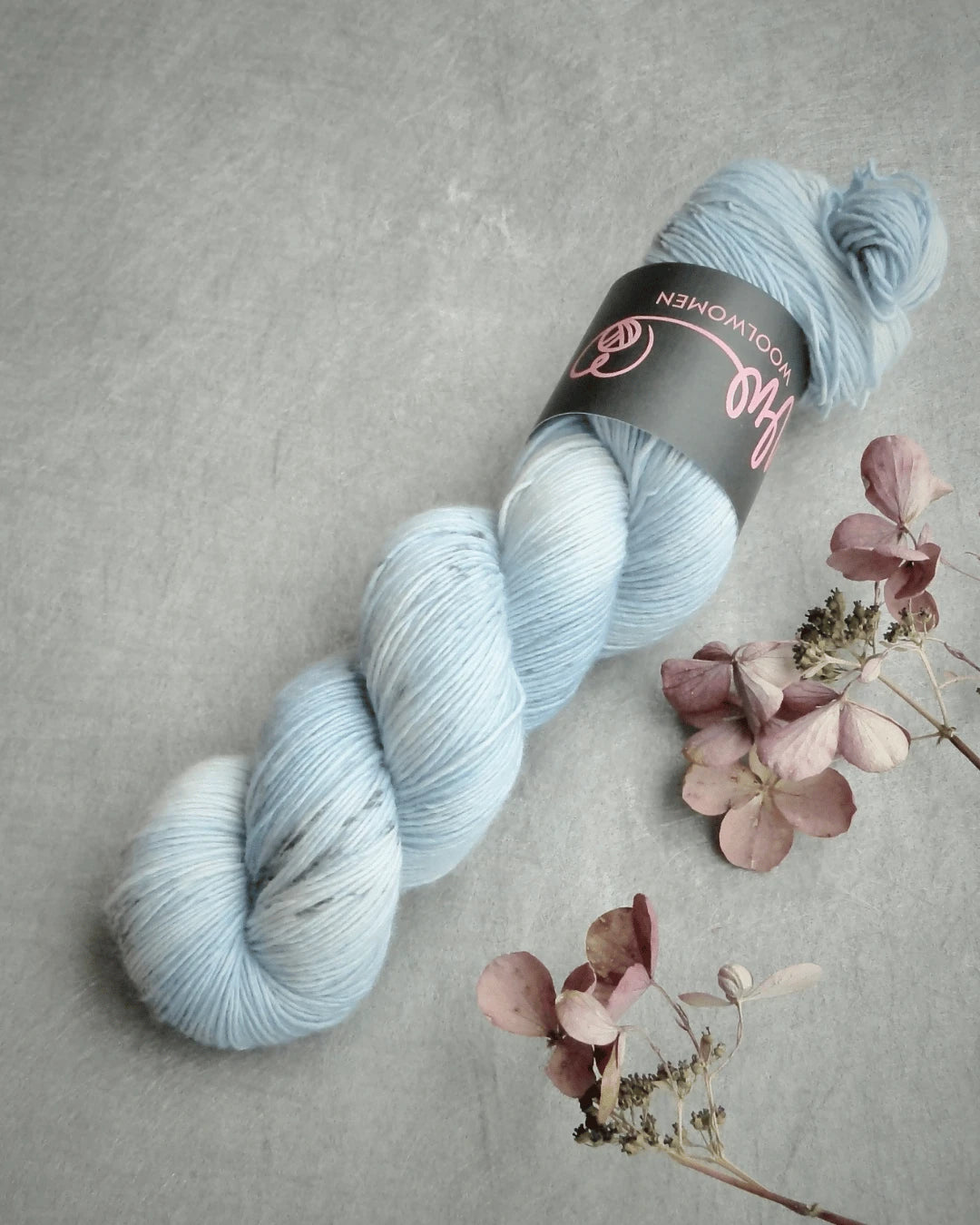 A skein of WoolWomen Merino Single, käsinvärjätty pale blue merinovilla yarn with subtle variegation and a black label, rests on a gray surface beside dried pink and purple hydrangea blooms.