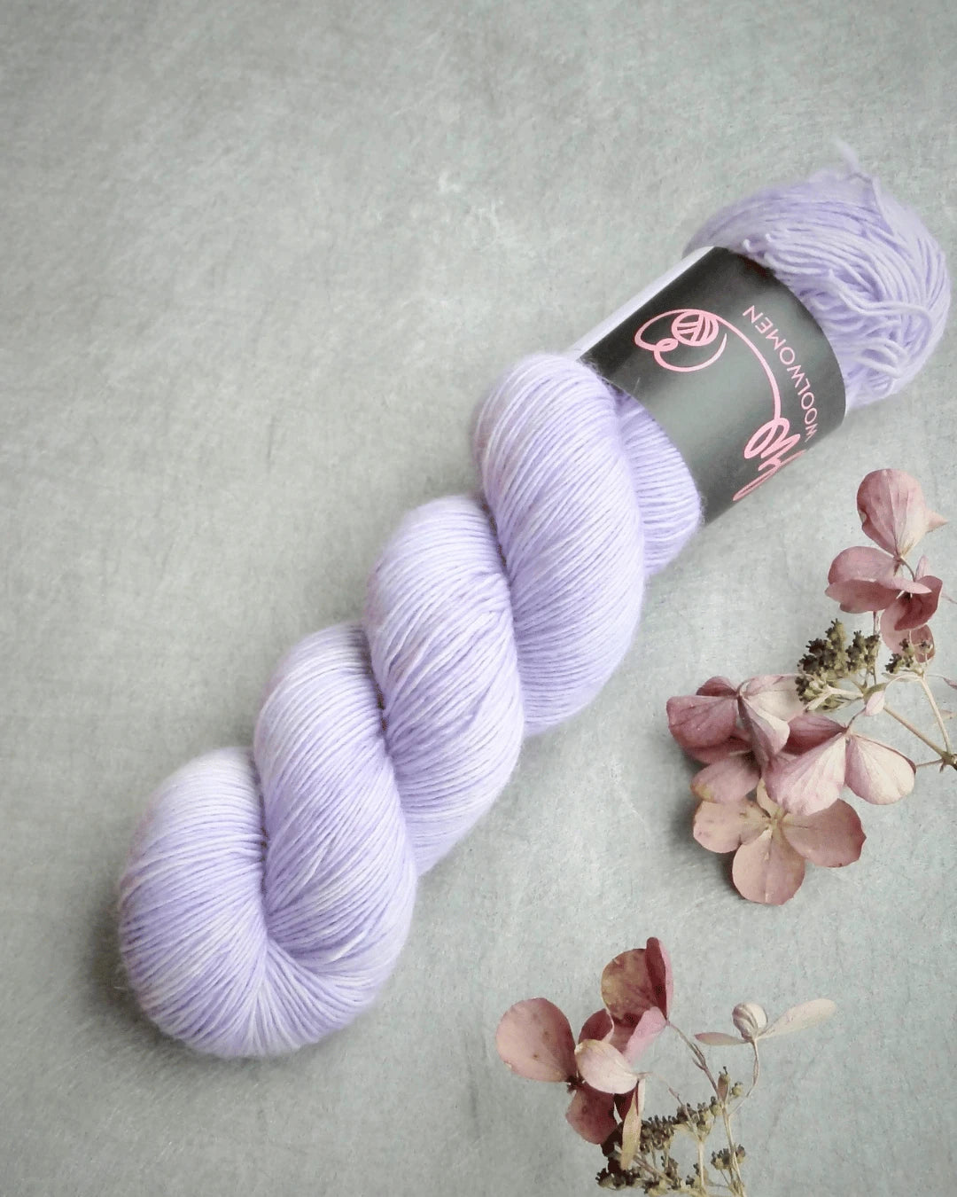 A skein of WoolWomen Merino Single soft, light purple yarn lies on a gray surface beside dried hydrangea flowers. The skein has a product label wrapped around it with a simple logo and text.