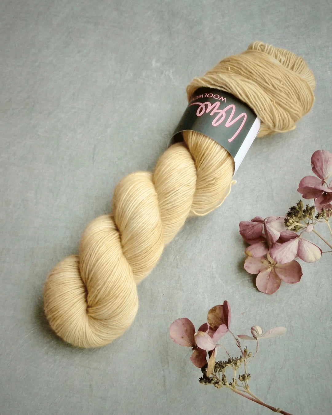 A skein of pale yellow WoolWomen Merino Single yarn with a black and pink label rests on a gray surface beside dried pink hydrangea flowers.