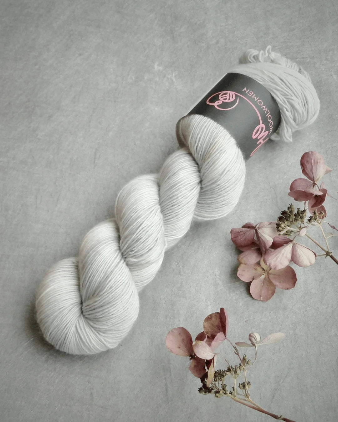 A skein of WoolWomen Merino Single yarn with a black label rests on a textured gray surface beside dried pink and brown hydrangea flowers.