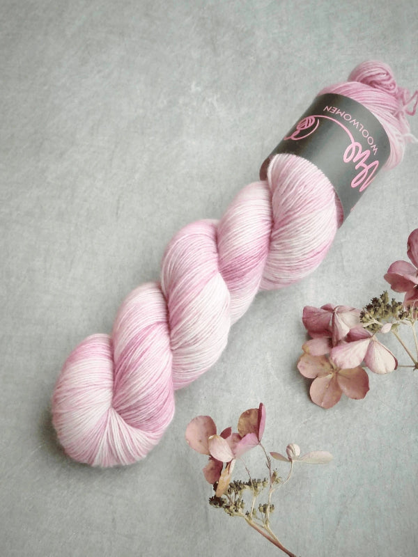 A skein of WoolWomen Merino Single in soft pink and white rests on a gray surface beside dried hydrangea flowers and petals.