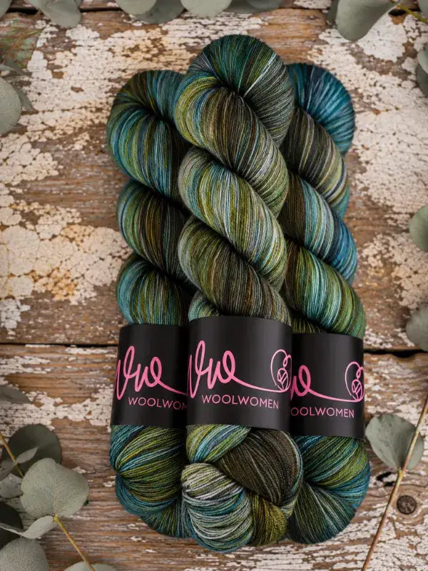 Three skeins of WoolWomen Merino Sock yarn in variegated shades of green, blue, and brown, held together by black paper bands.