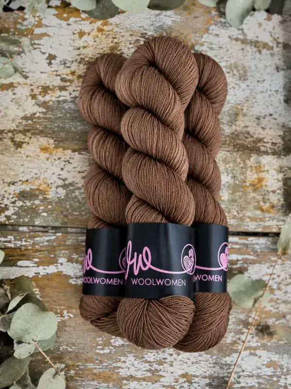 Three skeins of brown WoolWomen Merino Sock yarn wrapped in black branded paper bands.