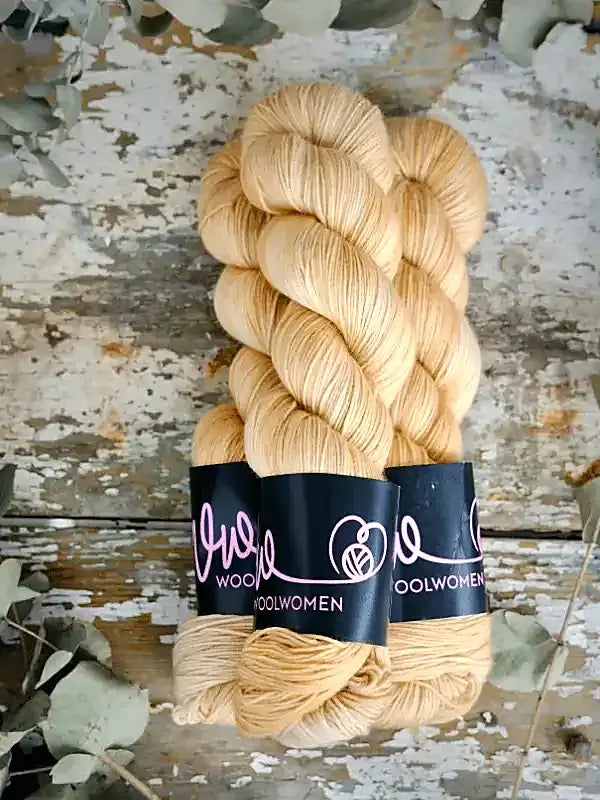 Two hanks of light golden-tan WoolWomen Merino Sock yarn with black labels, resting on a weathered wooden surface.